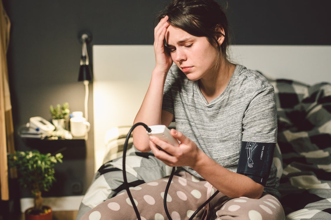 The topic of high blood pressure is hypertension disease. 22 year old young Caucasian woman uses an automatic tonometer to check pressure at home in the bedroom on the bed in home clothes in evening